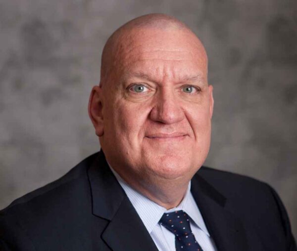 Ed Yohnka (a balk white man wearing a light blue collared shirt, dark blue tie with red and white dots, and a black suit jacket) poses in front of a gray backdrop