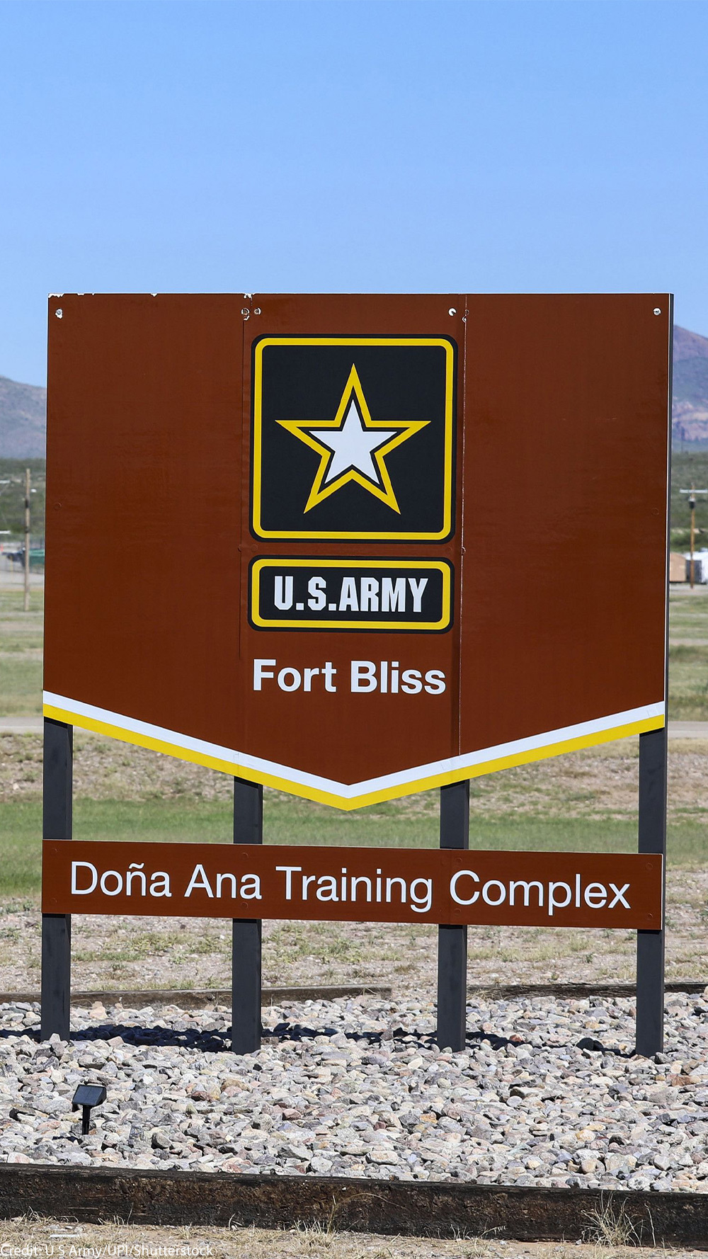 A large brown U.S. Army sign reading “Fort Bliss – Doña Ana Training Complex” stands in the foreground of a wide, open landscape. Behind it are low beige military buildings, utility poles, and a tall red-and-white checkered water tower, with mountains rising in the distance under a clear blue sky.