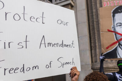 A person holds a large handwritten sign reading “Protect our First Amendment Freedom of Speech” during a protest outside the El Capitan Entertainment Centre, where Jimmy Kimmel Live! is filmed. Behind them, another protester in a skeleton costume holds a cardboard sign depicting a crossed-out image of Jimmy Kimmel. The demonstration is in response to ABC’s decision to pull Kimmel off the air following his remarks about Charlie Kirk.
