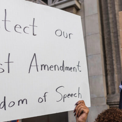 A person holds a large handwritten sign reading “Protect our First Amendment Freedom of Speech” during a protest outside the El Capitan Entertainment Centre, where Jimmy Kimmel Live! is filmed. Behind them, another protester in a skeleton costume holds a cardboard sign depicting a crossed-out image of Jimmy Kimmel. The demonstration is in response to ABC’s decision to pull Kimmel off the air following his remarks about Charlie Kirk.