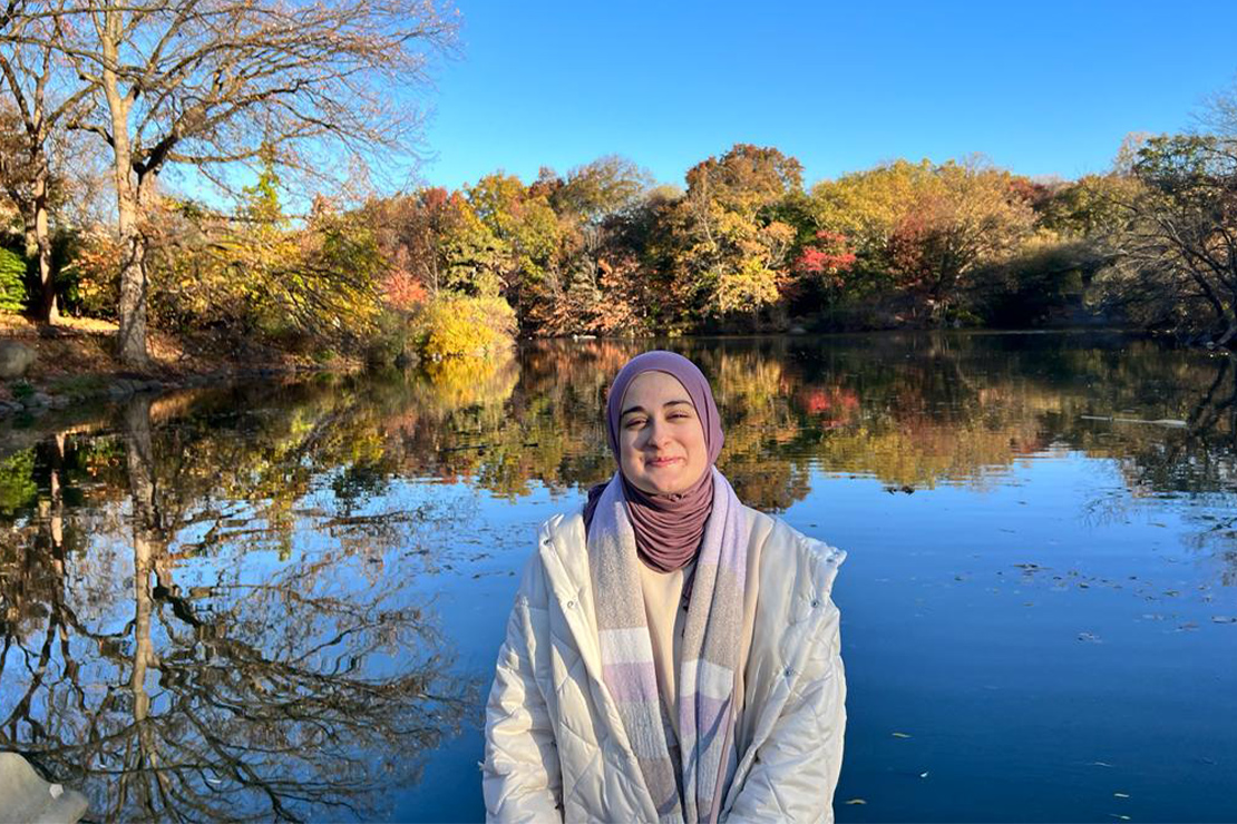 Rümeysa Öztürk in front of a lake during the fall with trees with browning leaves in the background.