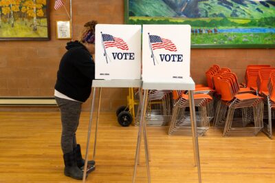 A person stands at a voting booth marked with an American flag and the word “VOTE” inside a community polling place. The room has wooden floors, orange stacked chairs, and colorful landscape paintings on the walls, with a small U.S. flag displayed nearby.