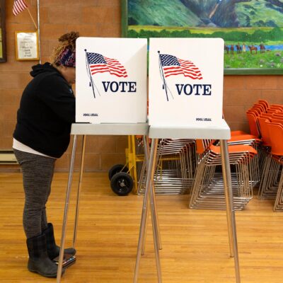 A person stands at a voting booth marked with an American flag and the word “VOTE” inside a community polling place. The room has wooden floors, orange stacked chairs, and colorful landscape paintings on the walls, with a small U.S. flag displayed nearby.